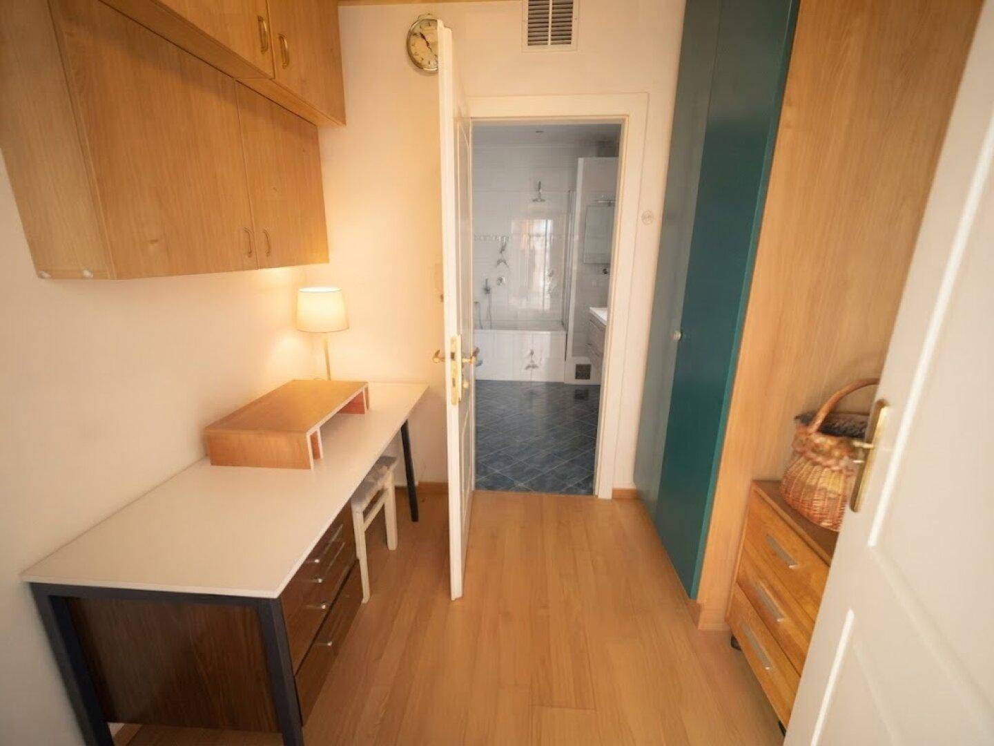 Kleines Arbeitszimmer mit Schreibtisch, Holzschränken und Blick ins Badezimmer.