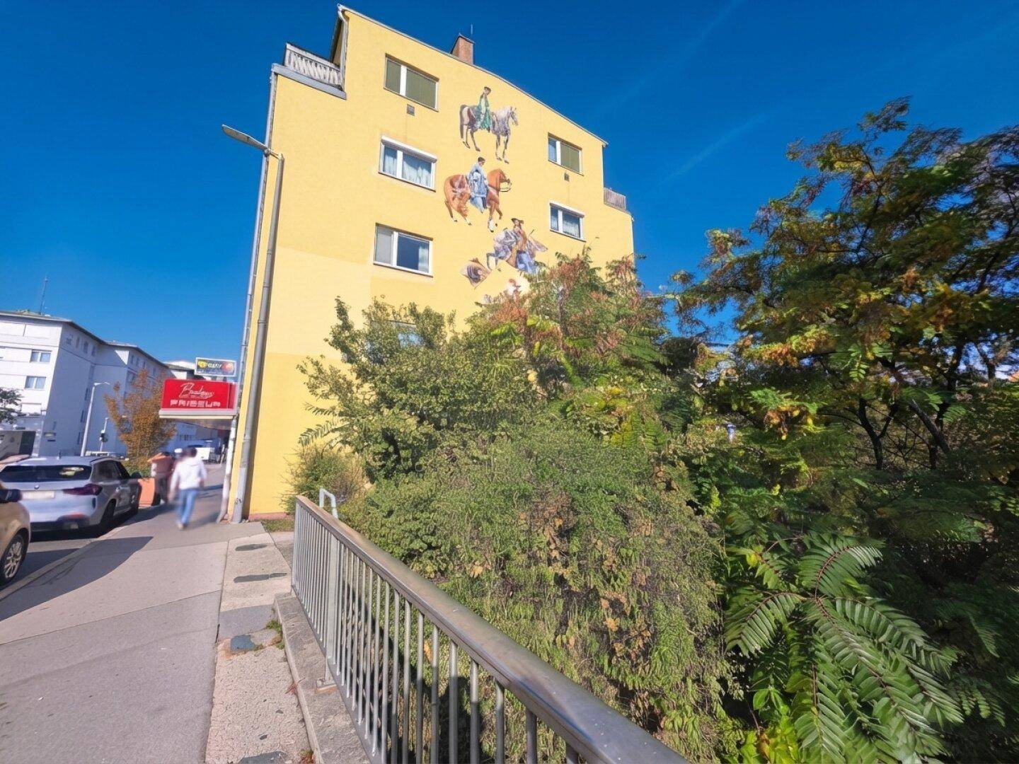 Gelbes Mehrfamilienhaus mit Wandgemälde und umliegender Vegetation.