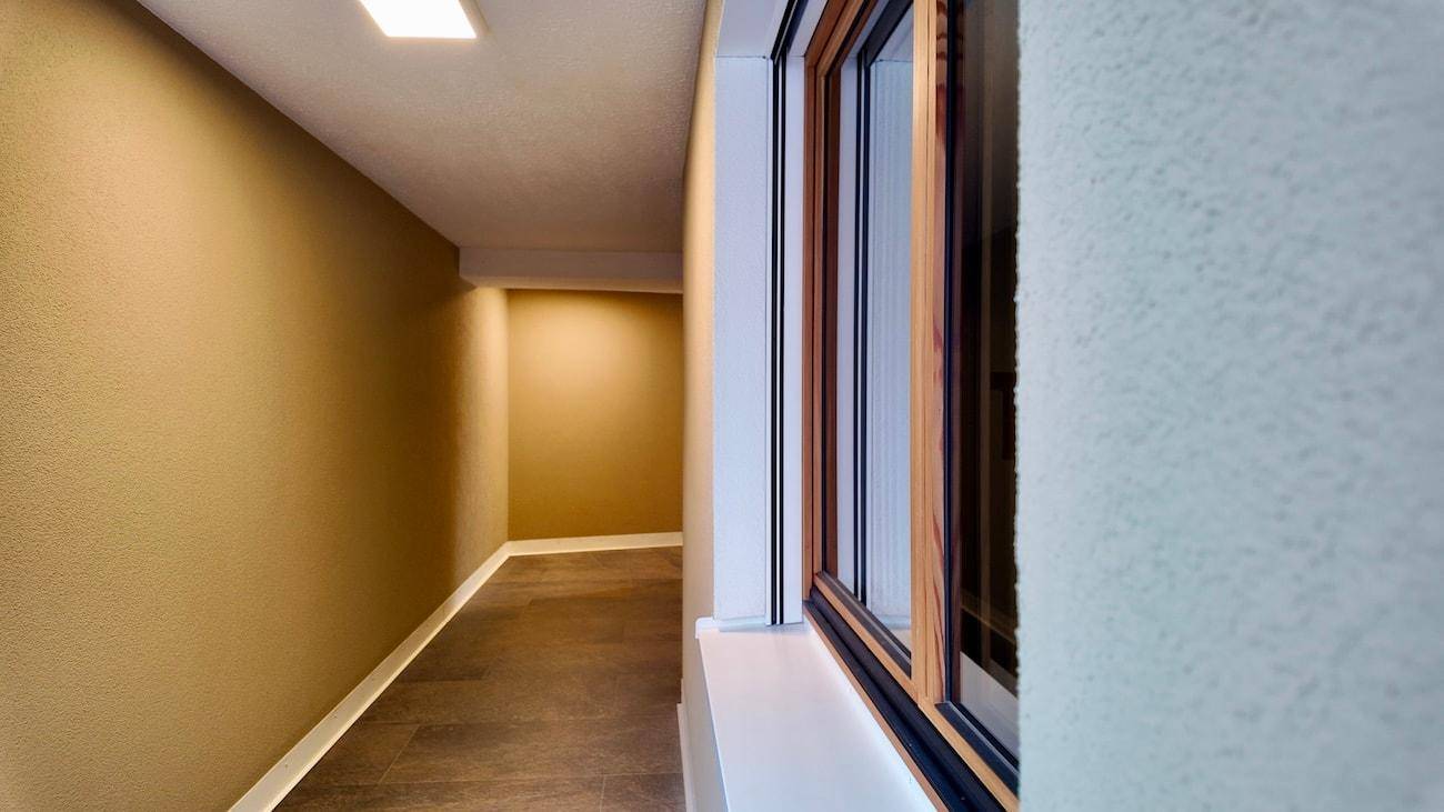 Heller Flur mit beigen Wänden, Fliesenboden und einem Fenster, das Tageslicht hereinlässt.