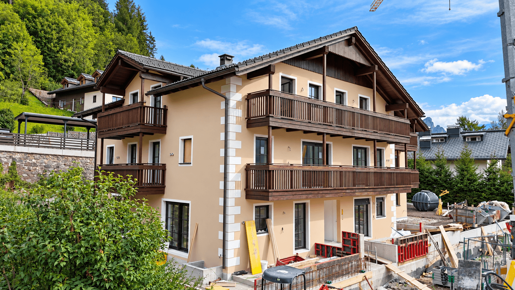Neubau im alpinen Stil mit Balkonen, umgeben von Baustelle und grüner Landschaft.