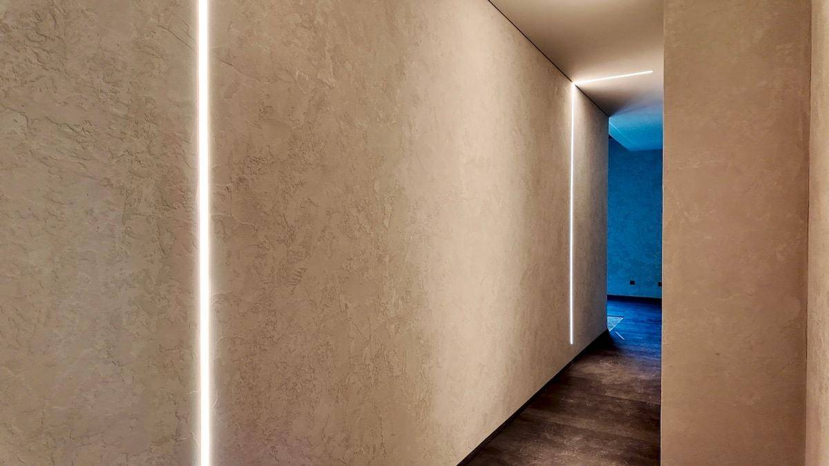 Langer Flur mit vertikalen Lichtleisten und strukturierten Wänden, die in einen blauen Raum führen.