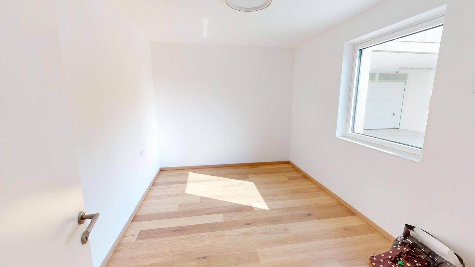 Leeres Zimmer mit hellem Holzboden und einem Fenster, das viel Tageslicht hereinlässt.