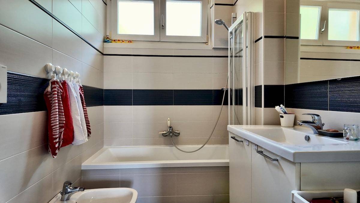 Helles Badezimmer mit Badewanne, Duschvorrichtung, WC und Bidet, gefliest in Grau und Blau.