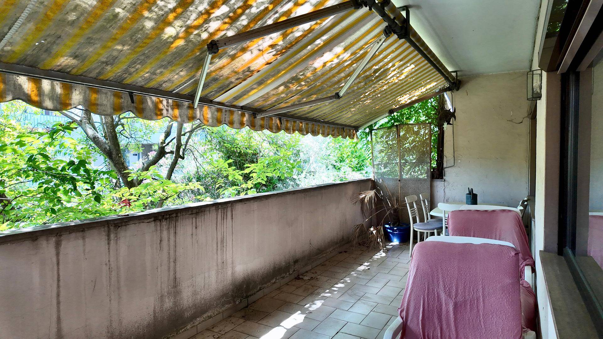 Geräumiger Balkon mit ausgefahrener Markise, Fliesenboden und Sitzgelegenheiten, umgeben von viel Grün.