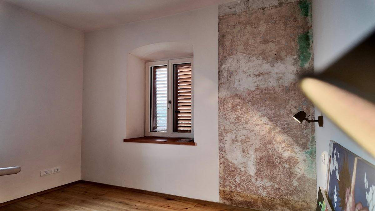 Raum mit Holzboden, Fenster mit Jalousien und einer Wand mit freigelegter Putzstruktur.