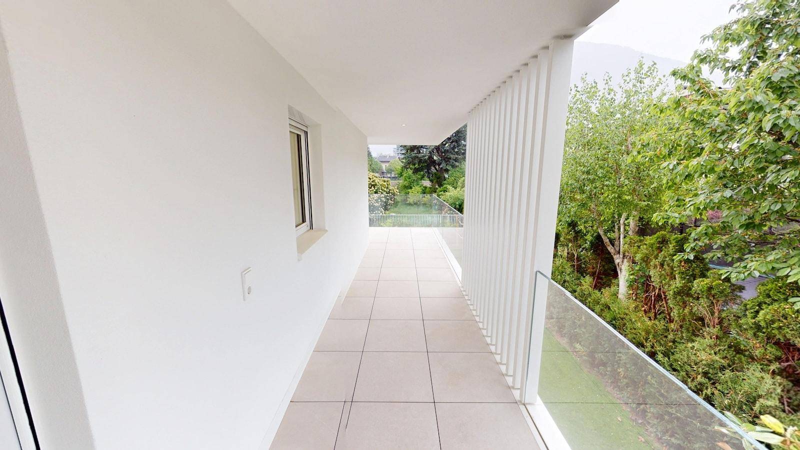 Moderner Balkon mit Fliesenboden und Glasgeländer, umgeben von üppigem Grün.