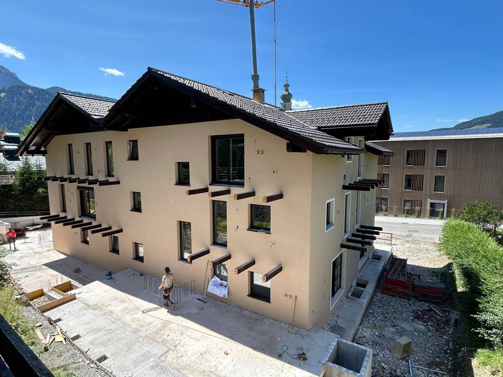Neubauhaus in alpiner Umgebung mit beiger Fassade und dunklem Dach, umgeben von Baustelle.