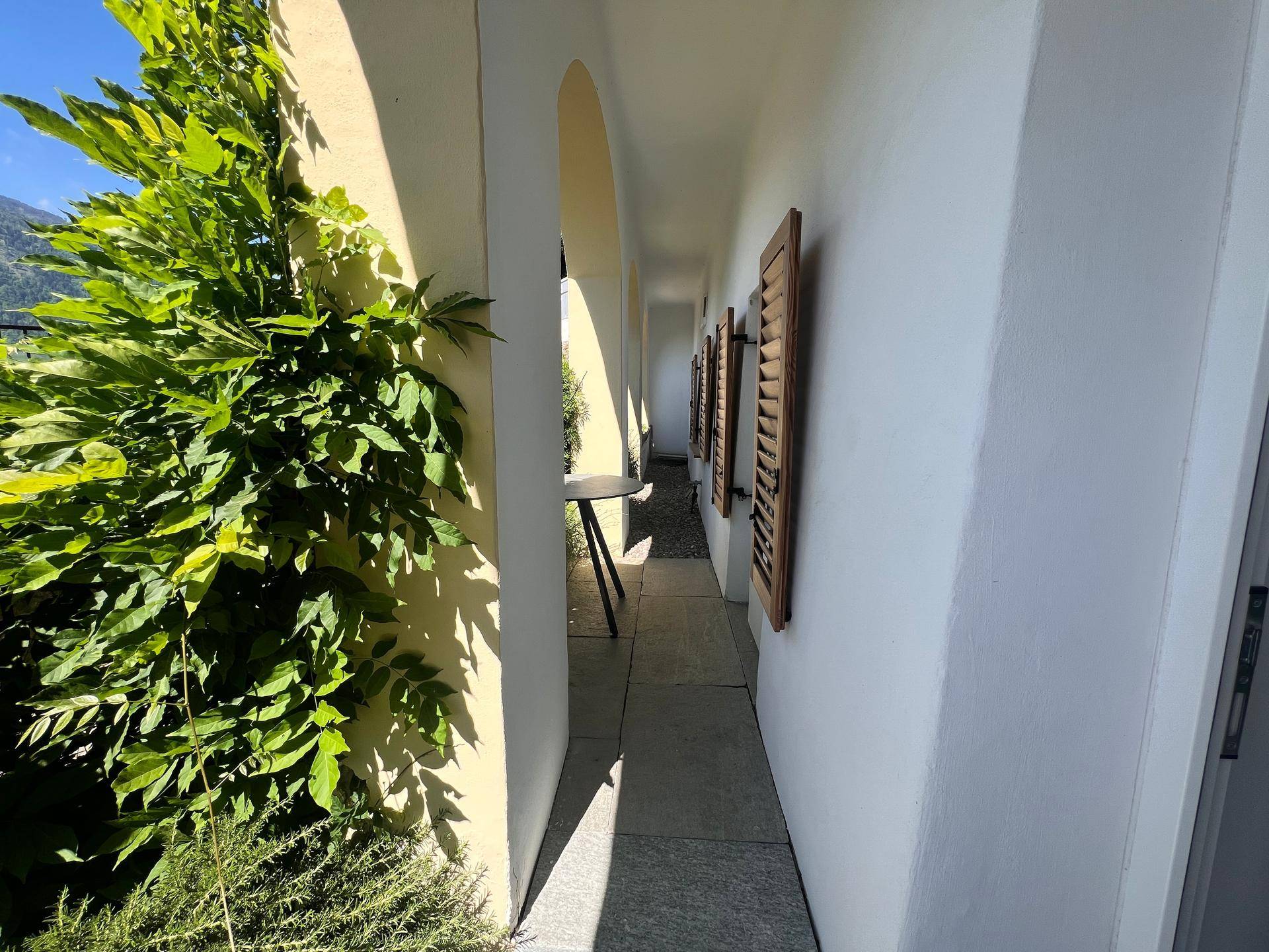 Langer, sonniger Außenkorridor mit Bögen, üppigem Grün und traditionellen Holzfensterläden.
