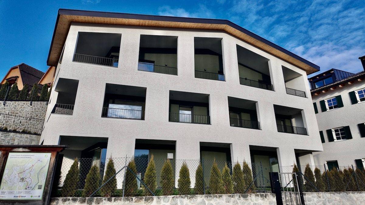 Modernes Wohngebäude mit weißer Fassade, dunklen Balkonen und gepflegten Hecken im Vordergrund.