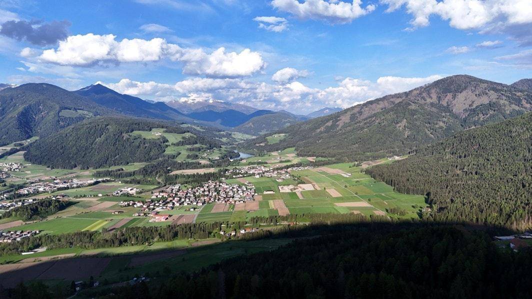 Panoramablick über ein grünes Tal mit einem Fluss, umgeben von bewaldeten Hügeln und schneebedeckten Bergen.