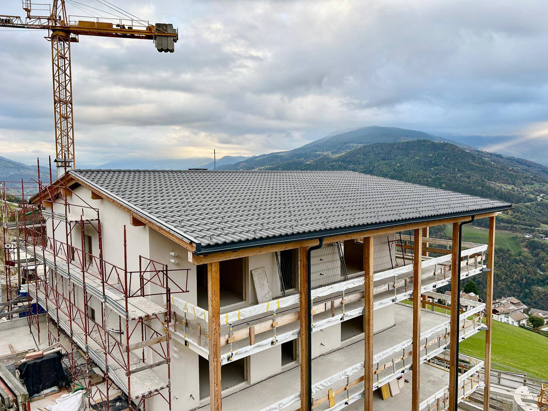 Neubau im Bau mit Gerüst und Dachziegeln, umgeben von einer malerischen Berglandschaft.
