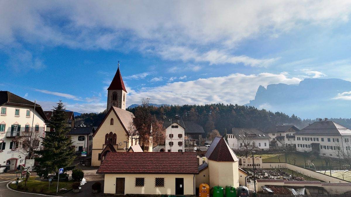 Panoramablick auf ein malerisches Dorf mit Kirche und Bergen im Hintergrund unter blauem Himmel.