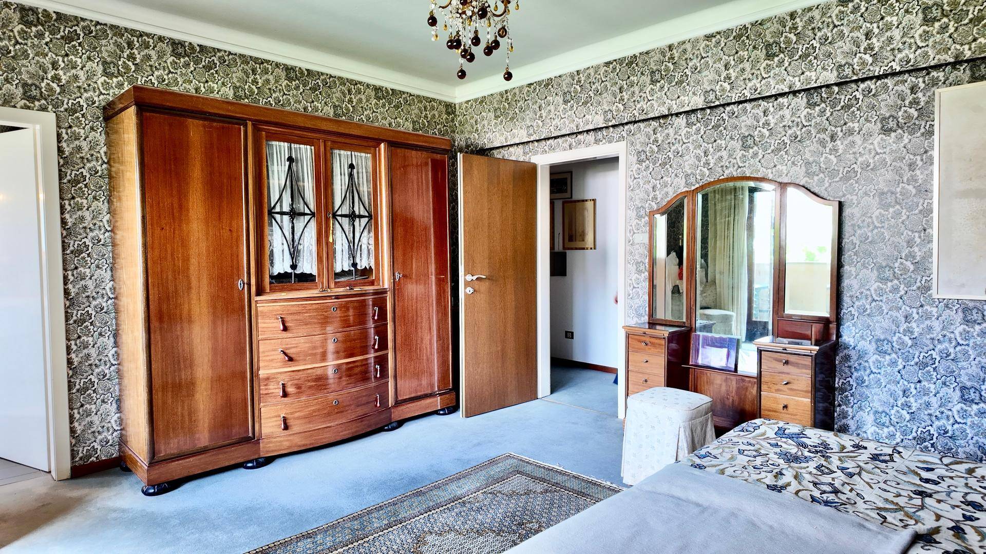 Schlafzimmer mit großem Holzschrank, Schminktisch und gemusterter Tapete.