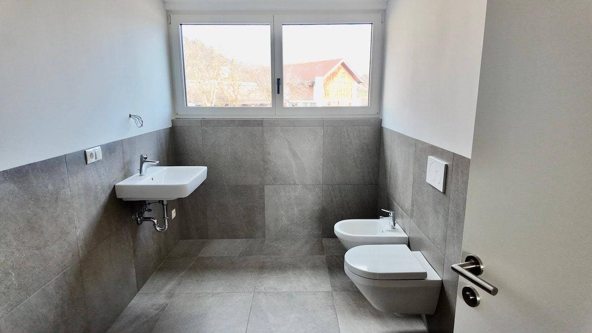 Zeitgemäßes Badezimmer mit grauen Fliesen, Bidet, wandhängendem WC und Fenster.