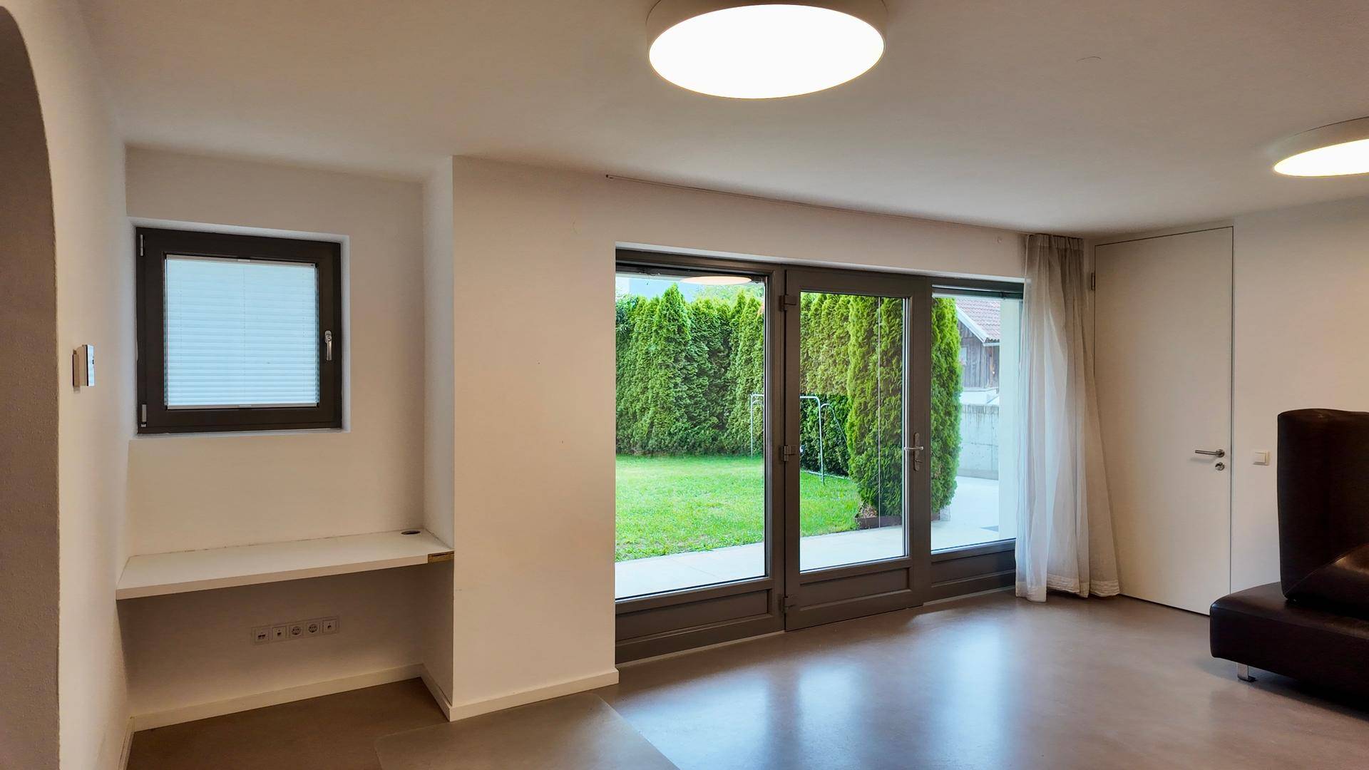 Geräumiger Wohnbereich mit großen Fenstern zum Garten und einer praktischen Ablage unter einem kleineren Fenster.
