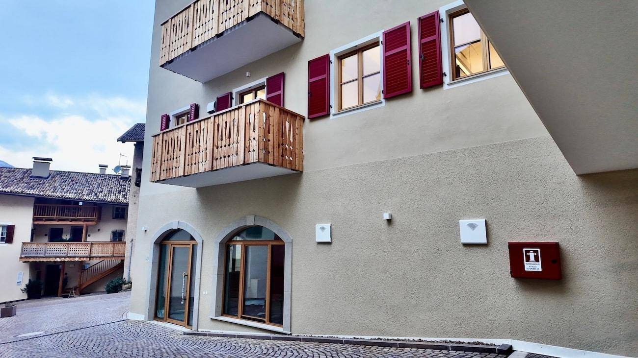 Gebäudefassade mit roten Fensterläden, Holzbalkonen und einem gepflasterten Eingangsbereich.