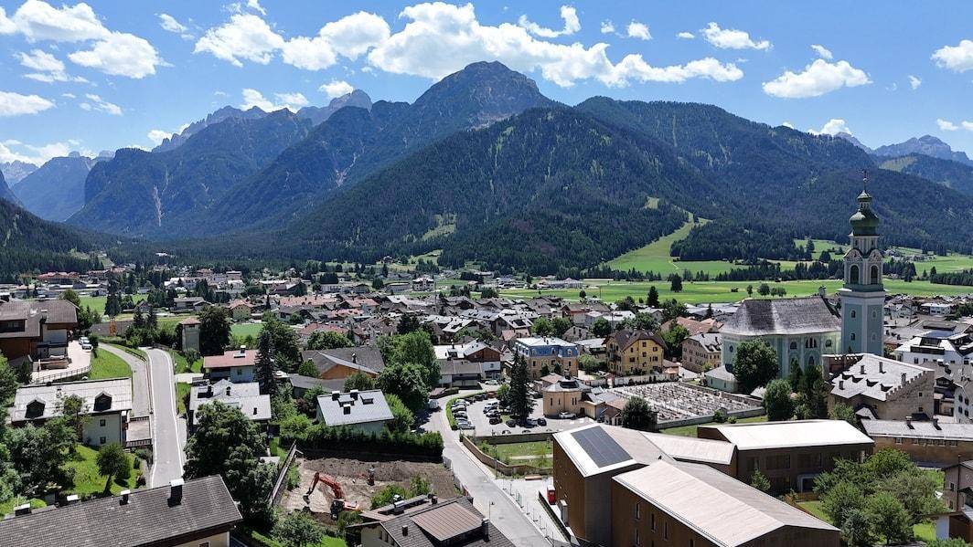 Panoramablick auf eine malerische Stadt in den Alpen, umgeben von grünen Wiesen und hohen Bergen.