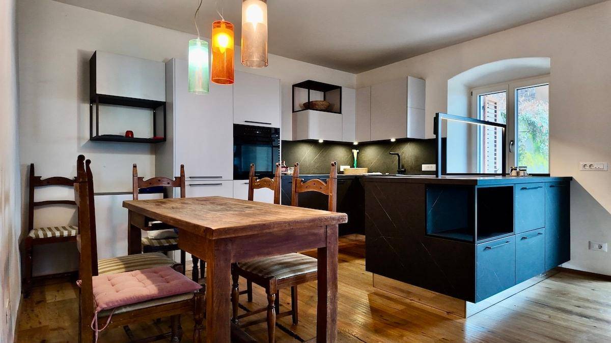 Moderne Wohnküche mit dunkler Kücheninsel, Esstisch und bunten Hängelampen.