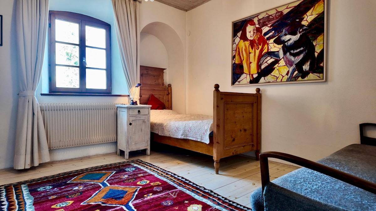 Helles Schlafzimmer mit Einzelbett aus Holz, gemustertem Teppich und einem Fenster mit Blick nach draußen.