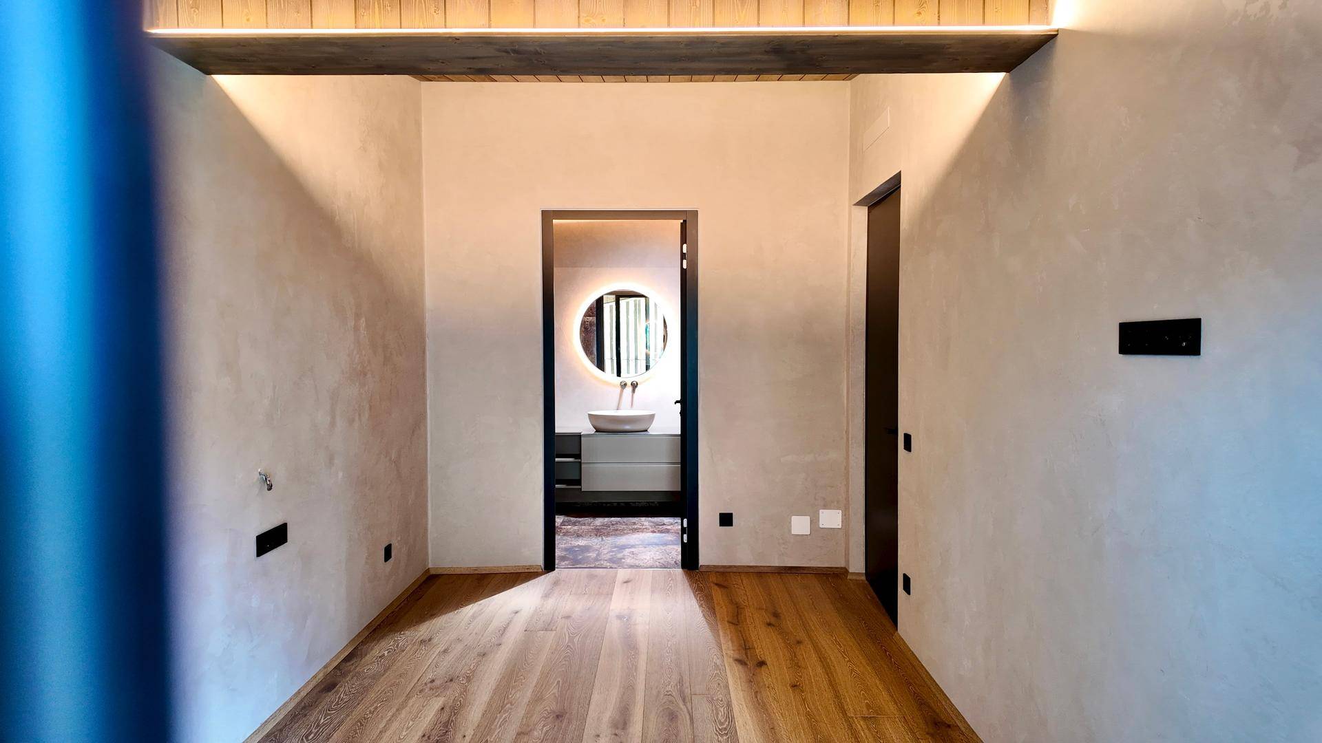 Schlafzimmer mit Holzboden und Blick in ein modernes Bad mit rundem Spiegel.