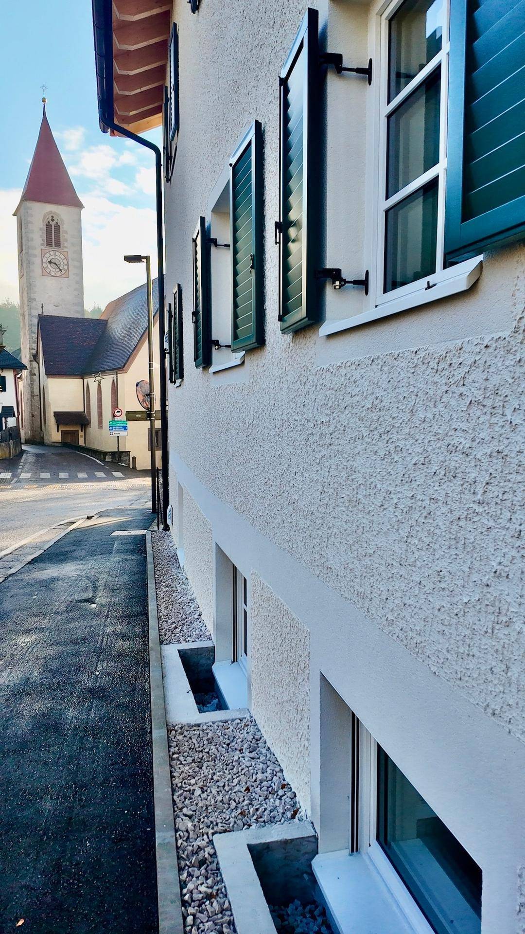 Seitenansicht eines Gebäudes mit grünen Fensterläden und einem Kirchturm in der Ferne.