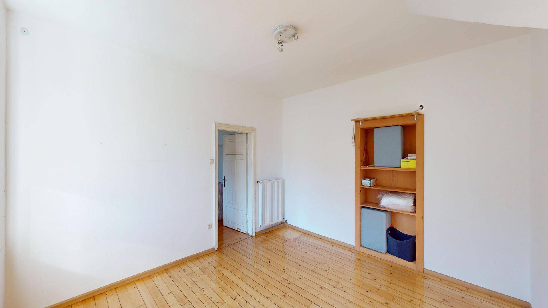 Leeres Zimmer mit Holzdielenboden, Heizkörper und eingebautem Regal.