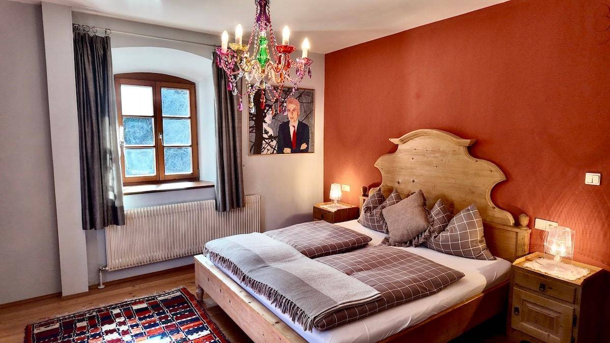 Gemütliches Schlafzimmer mit rustikalem Holzdoppelbett, farbenfrohem Kronleuchter und einem Fenster mit Heizkörper.