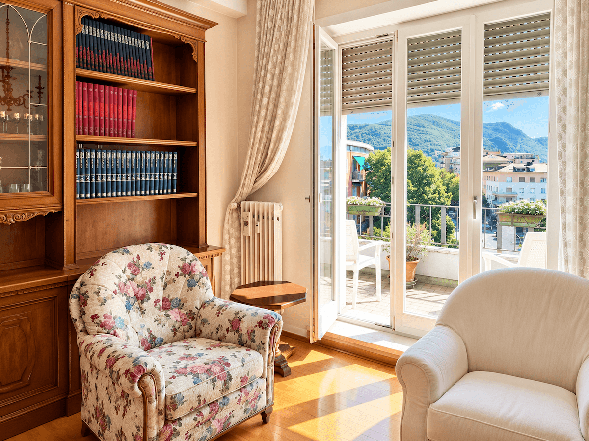 Gemütliches Wohnzimmer mit klassischem Holzregal, floralem Sessel und Zugang zum sonnigen Balkon mit Bergblick.