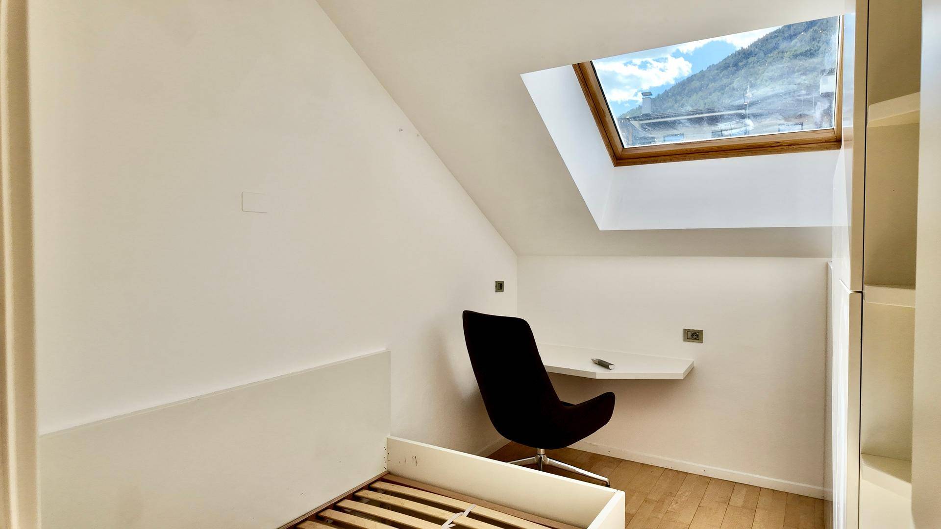 Helles Zimmer im Dachgeschoss mit Schreibtisch, Bettrahmen und Dachfenster.