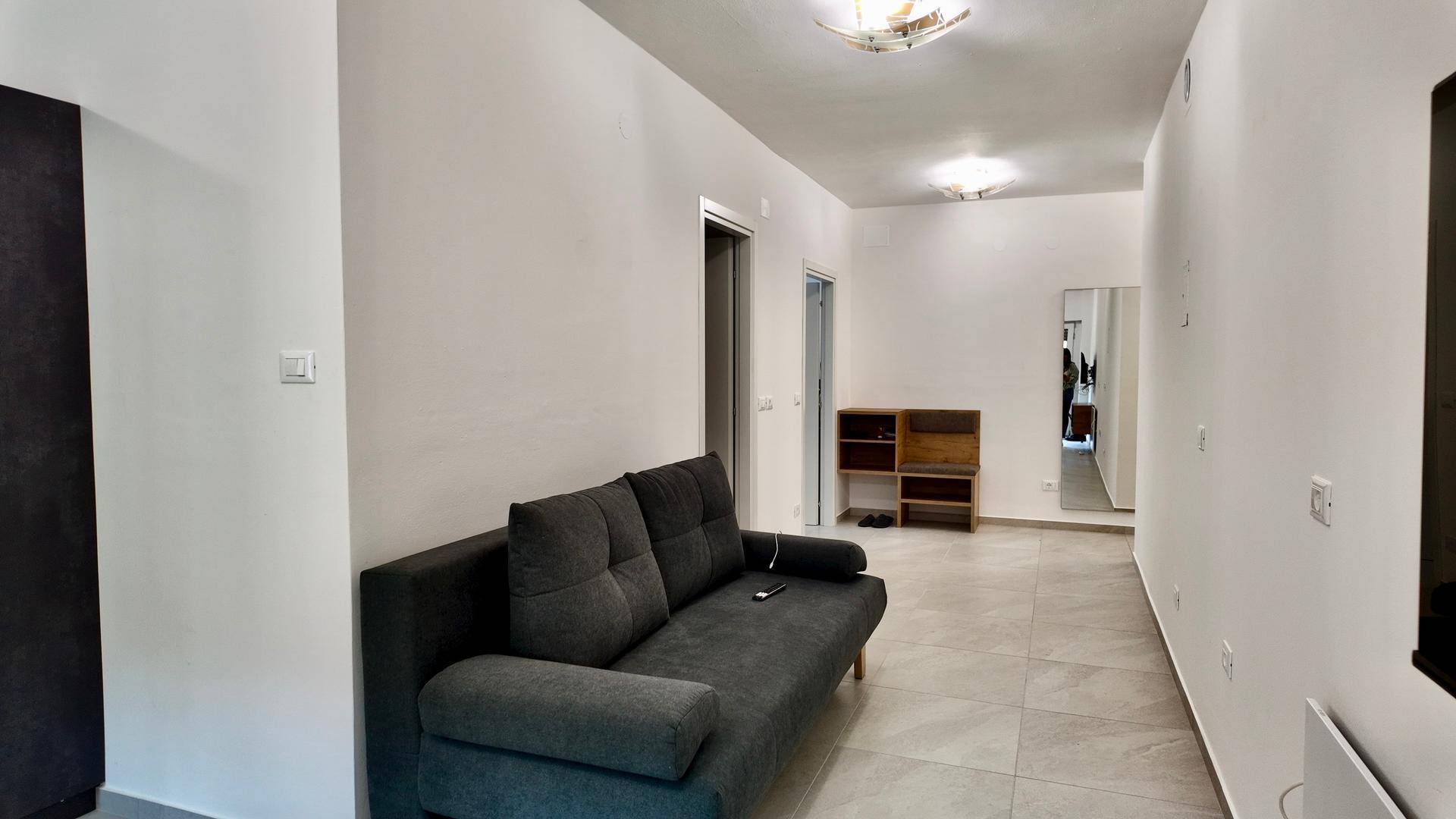 Heller Wohnbereich mit grauem Sofa, Fliesenboden und einem Blick in den Flur mit Spiegel.