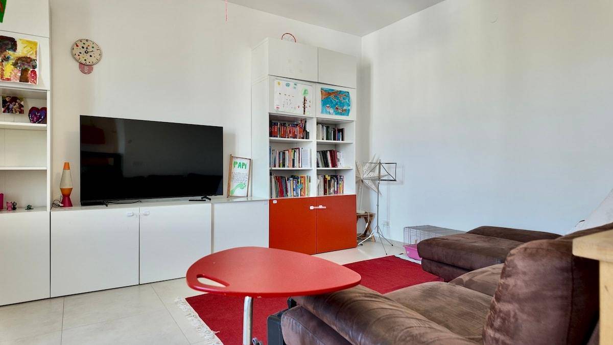 Gemütliches Wohnzimmer mit großem Fernseher, Bücherregal und bequemer Couch auf rotem Teppich.