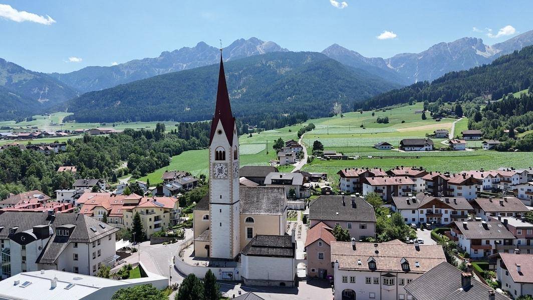 Luftaufnahme eines malerischen Dorfes mit Kirchturm, umgeben von grünen Feldern und Bergen.