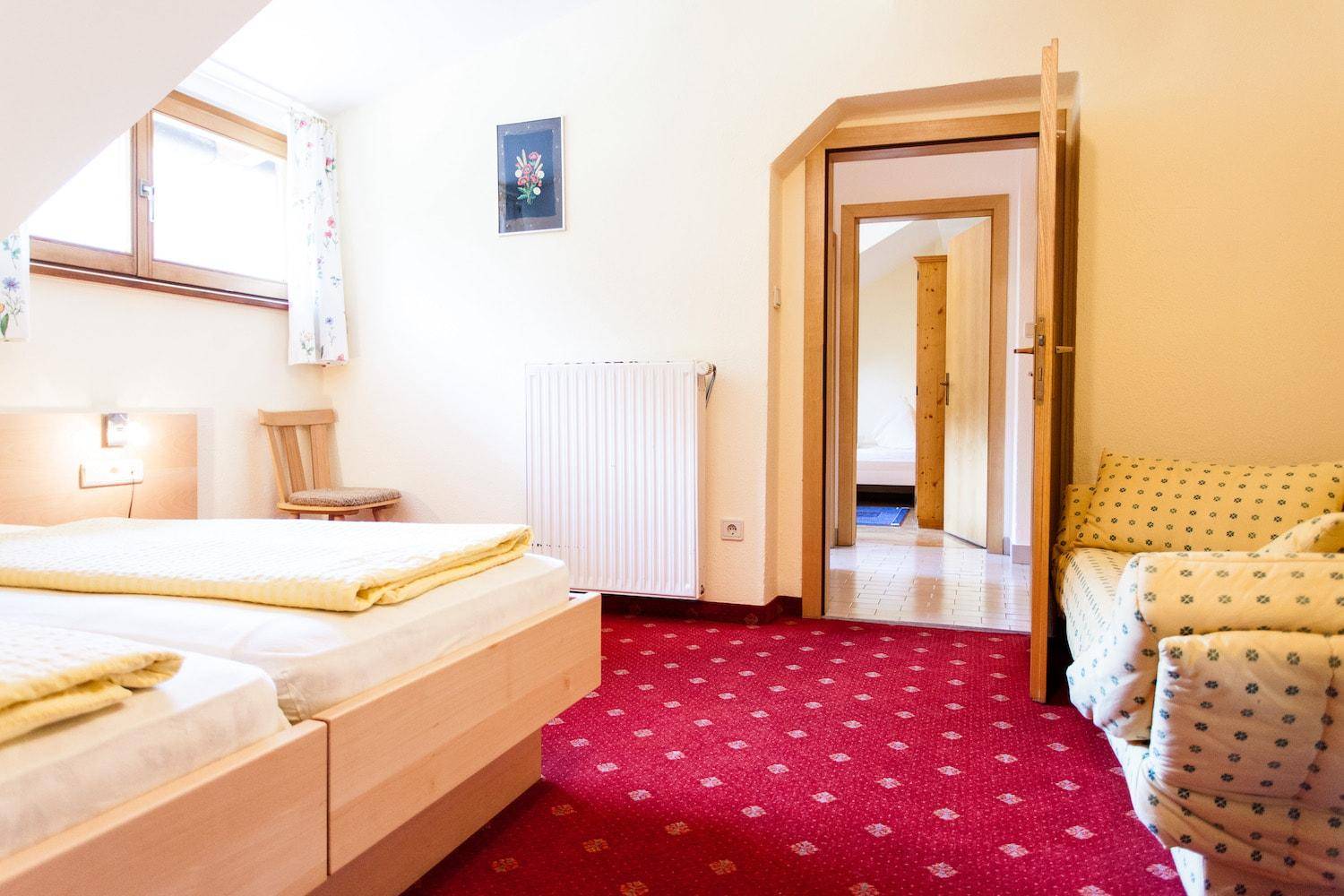 Helles Schlafzimmer mit rotem Teppichboden, Fenster und Zugang zu einem weiteren Raum.