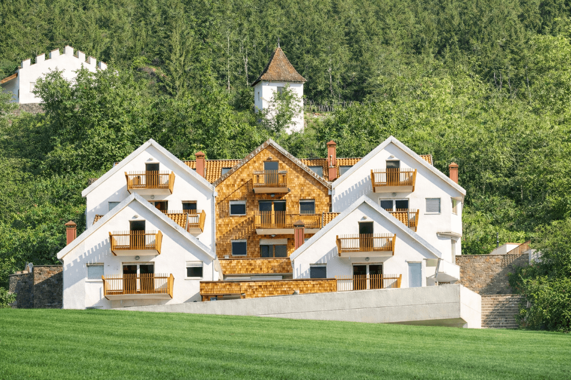 Mehrere Gebäude mit Holzbalkonen und grüner Umgebung vor einem bewaldeten Hügel.