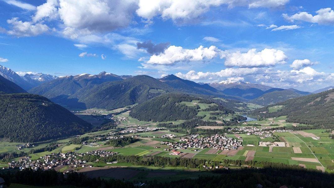 Weitläufige Berglandschaft mit grünen Tälern, bewaldeten Hügeln und kleinen Dörfern unter blauem Himmel.