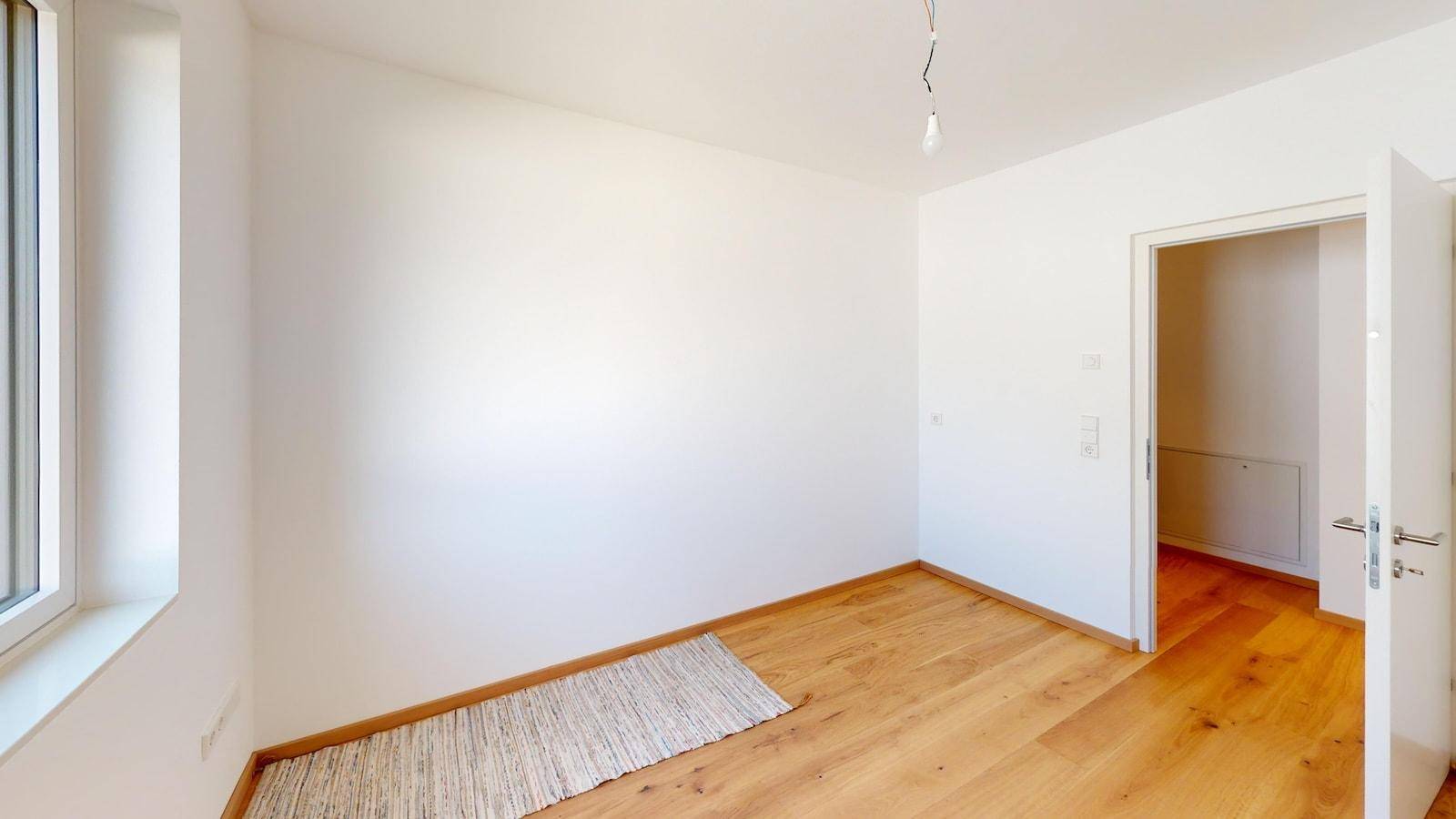 Helles, leeres Zimmer mit Holzboden und einem Fenster, das viel Tageslicht hereinlässt.
