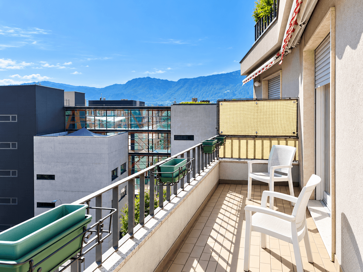 Sonniger Balkon mit Sitzgelegenheiten und Blumenkästen, mit Blick auf moderne Gebäude und die Bergkette.