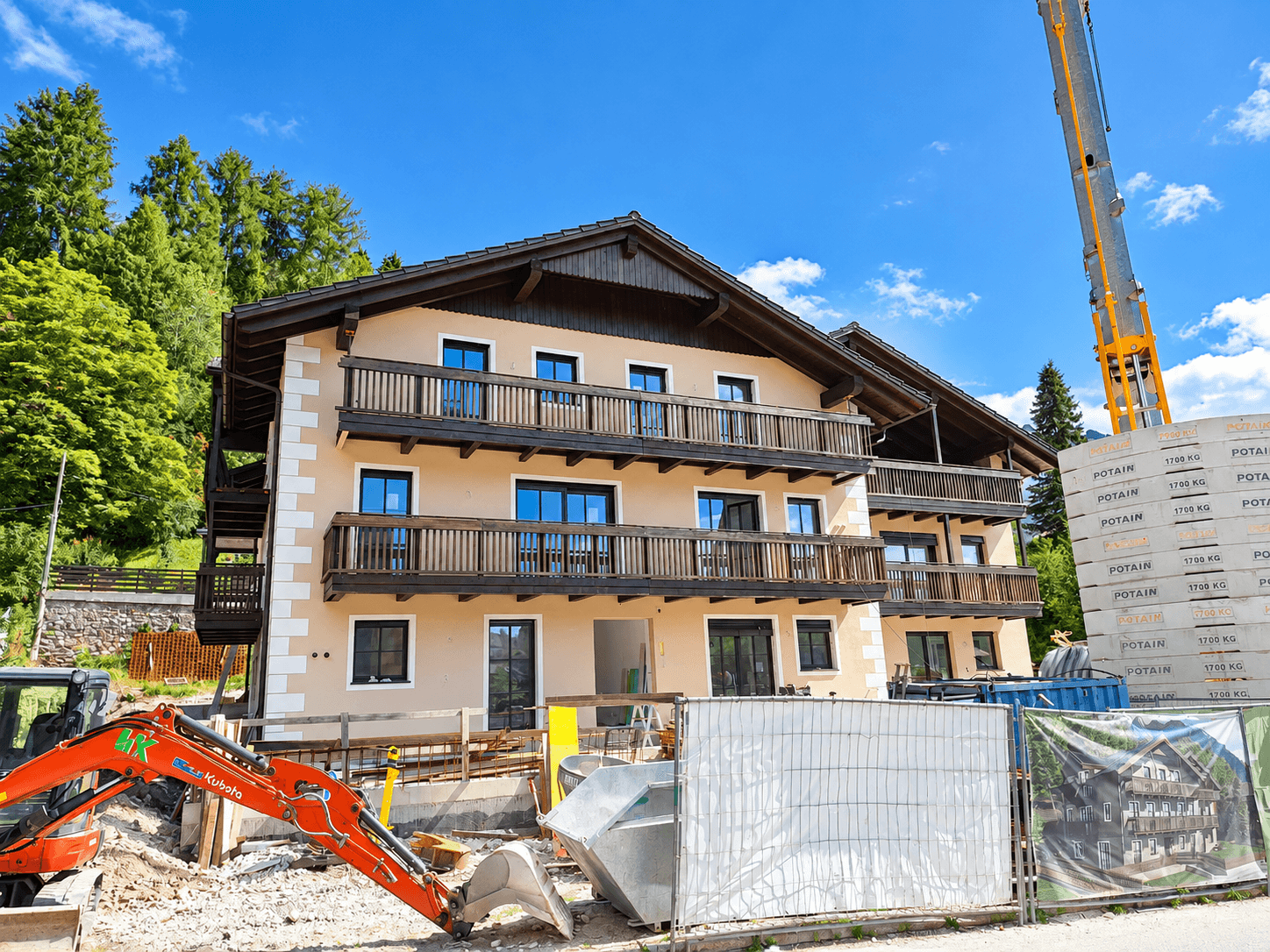 Neubau im alpinen Stil mit Balkonen, umgeben von Baustelle und grüner Berglandschaft.