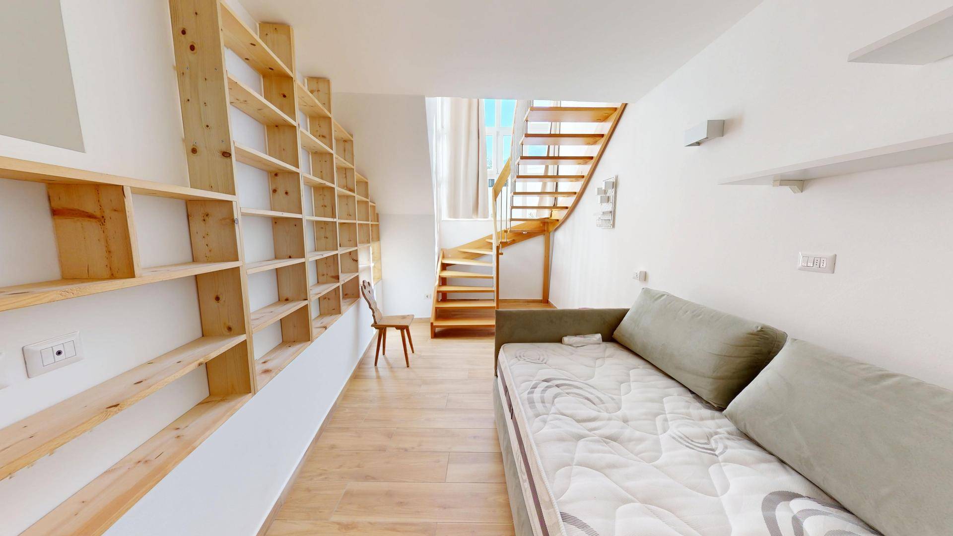 Helles Zimmer mit Schlafsofa, Holzregalen und einer Treppe, die zu einer oberen Ebene führt.