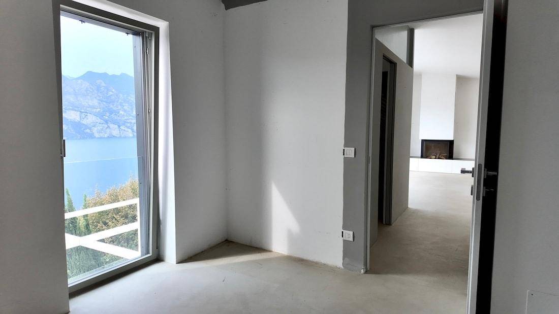 Leerer Raum mit Betonboden und weiß gestrichenen Wänden, mit Zugang zu einem Balkon und Seeblick.