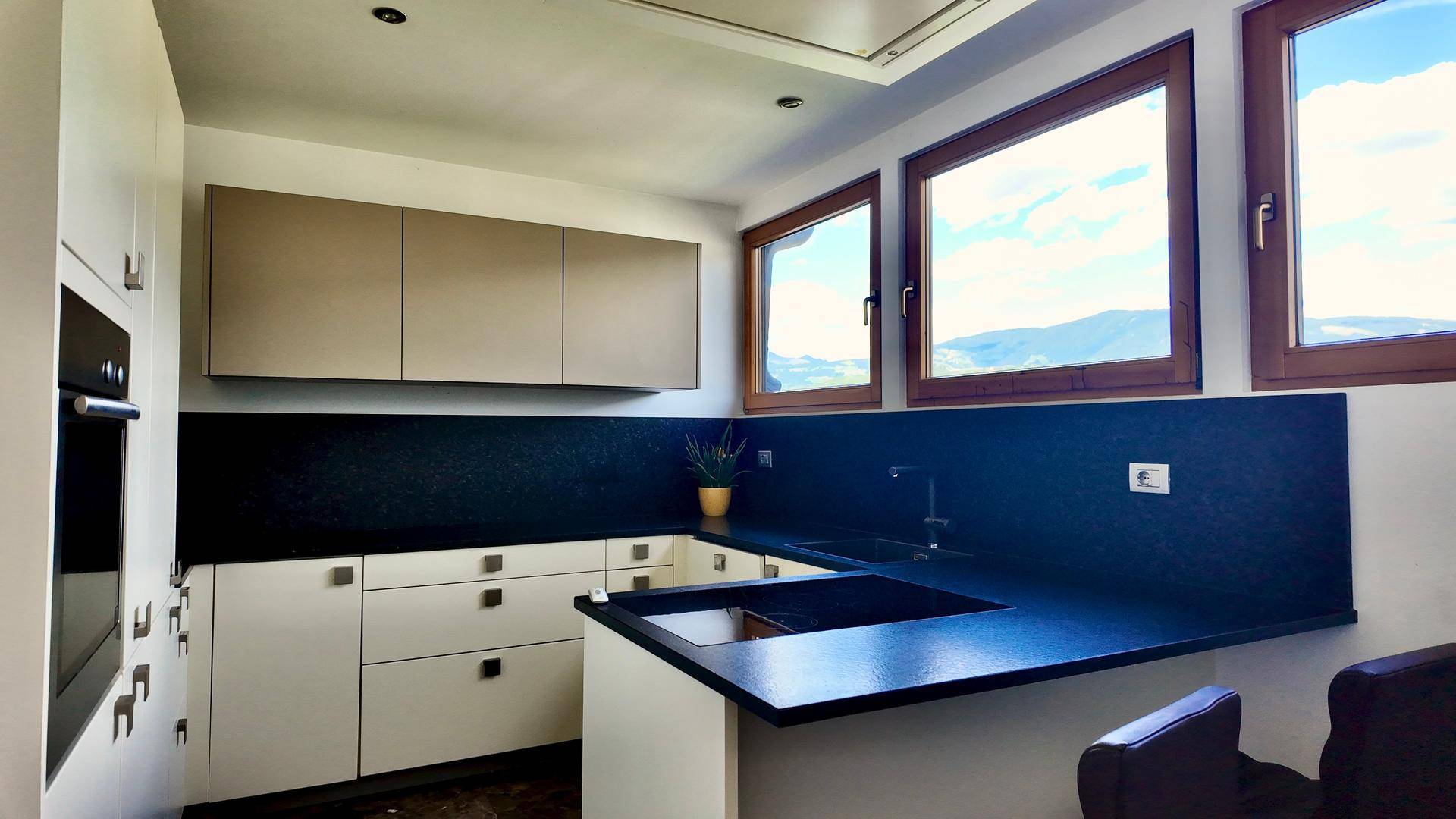 Moderne Küche mit dunklen Arbeitsplatten, weißen Schränken und Bergblick durch die Fenster.