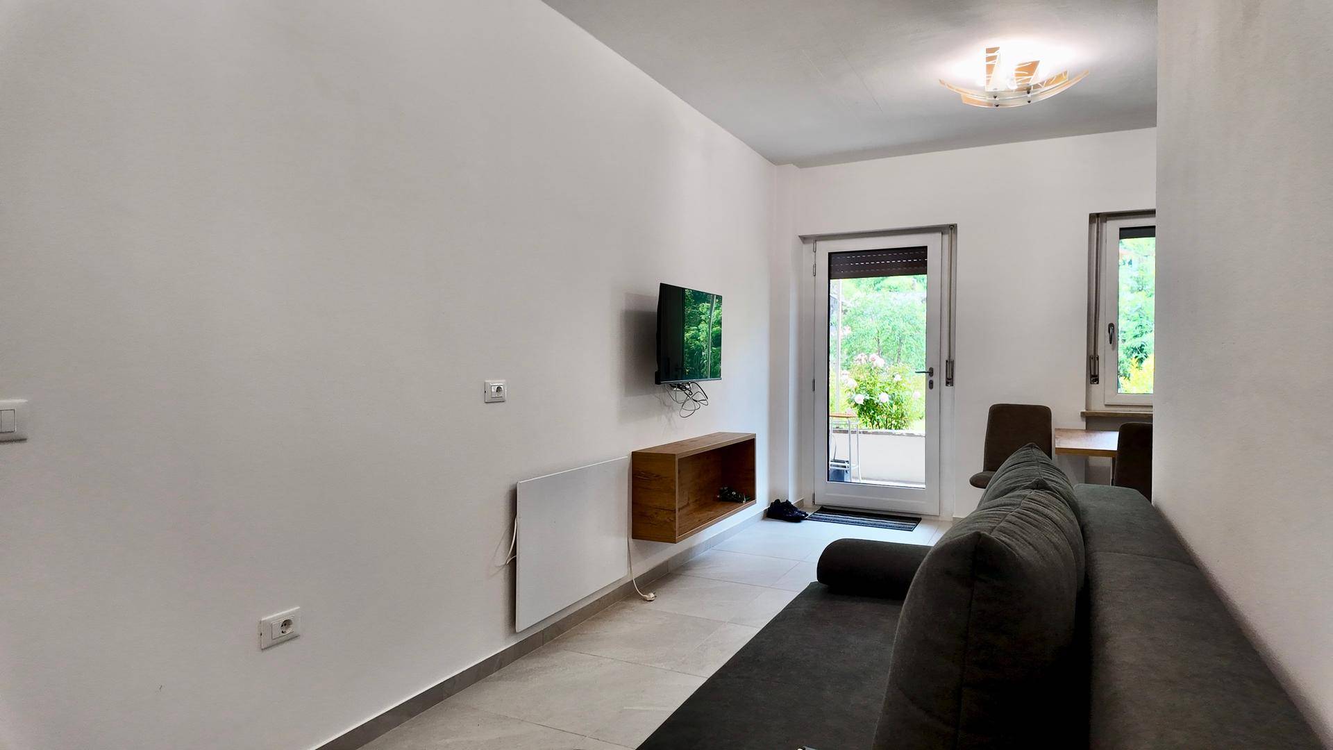 Wohnzimmer mit bequemem grauem Sofa, Wandfernseher und Zugang zu einem Außenbereich.