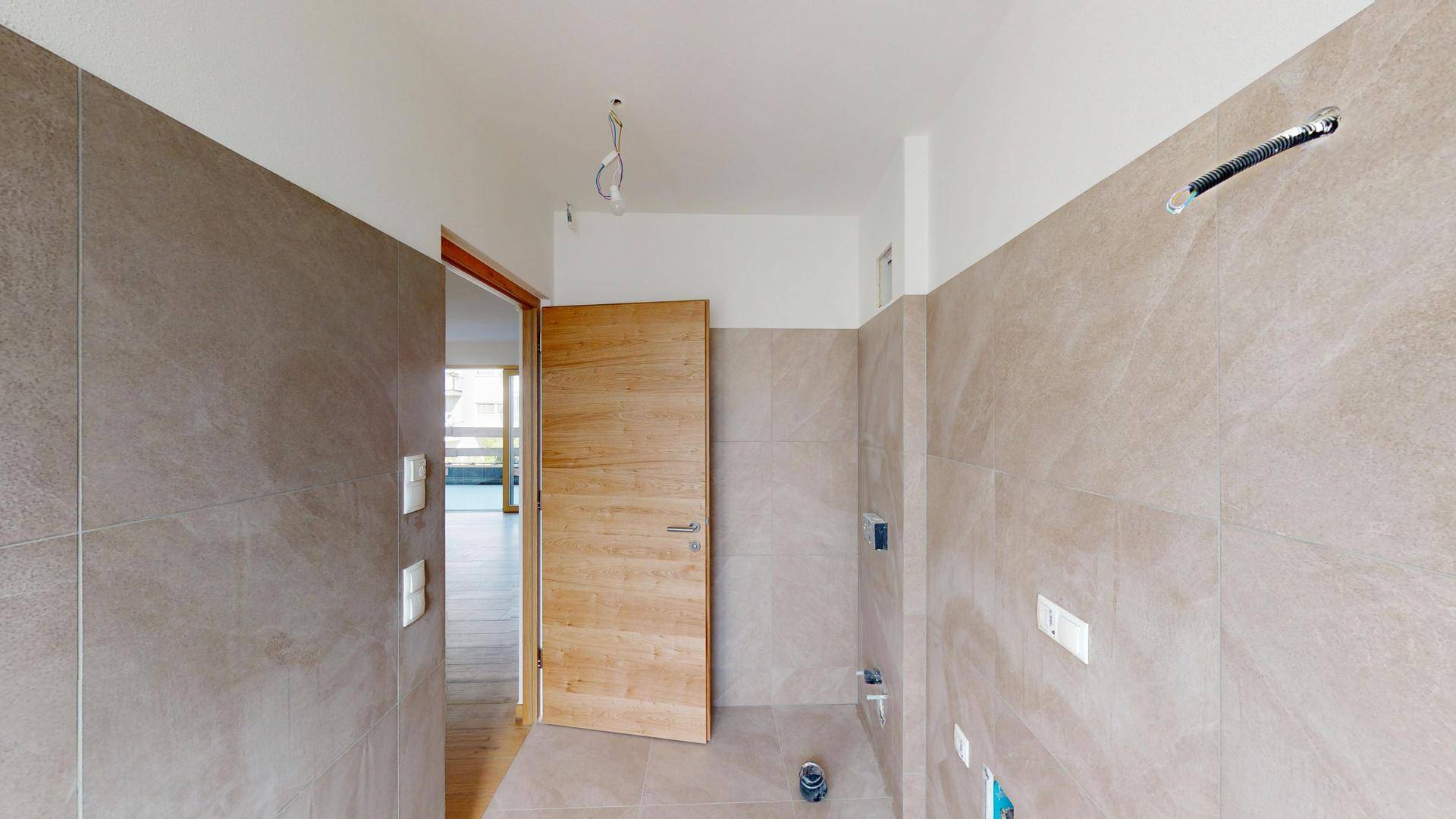 Geräumiges, unfertiges Badezimmer mit beigefarbenen Fliesen und einer Holztür.