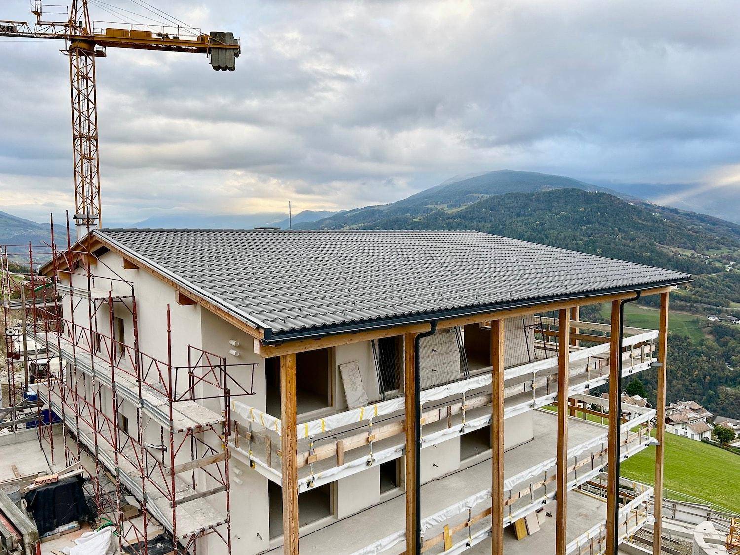 Im Bau befindliches Gebäude mit neu gedecktem Dach und Holzverkleidung an den Balkonen, umgeben von grünen Bergen.