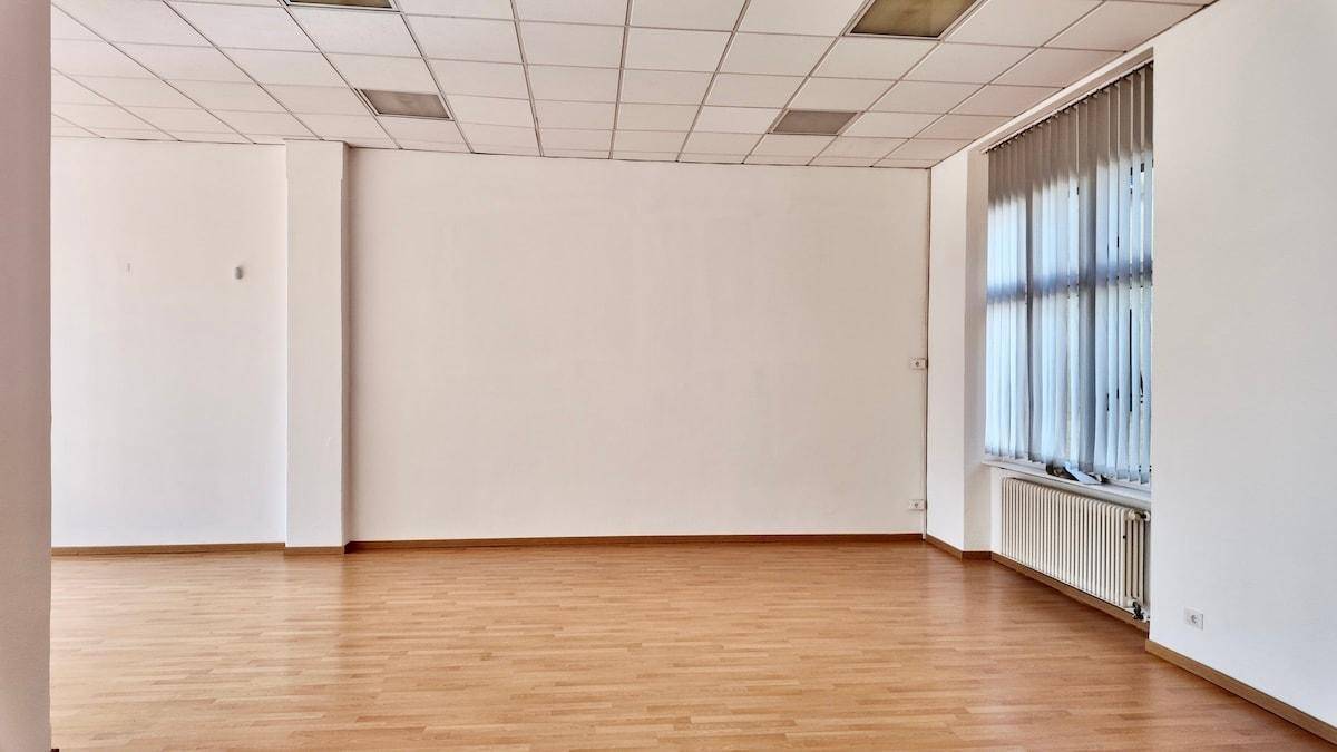 Heller, leerer Büroraum mit Holzfußboden, weißen Wänden und einem Fenster mit Jalousien.