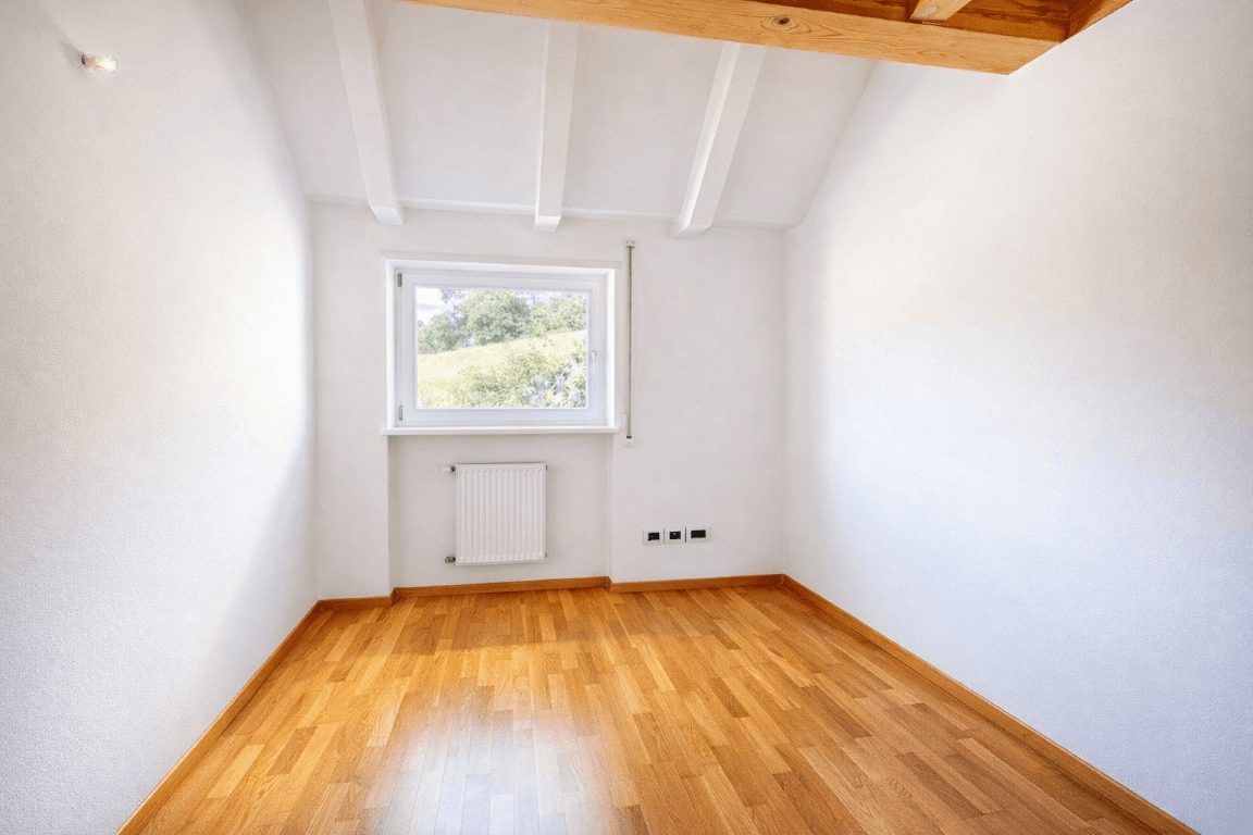Heller, leerer Raum mit Holzboden und Fenster mit Blick ins Grüne.