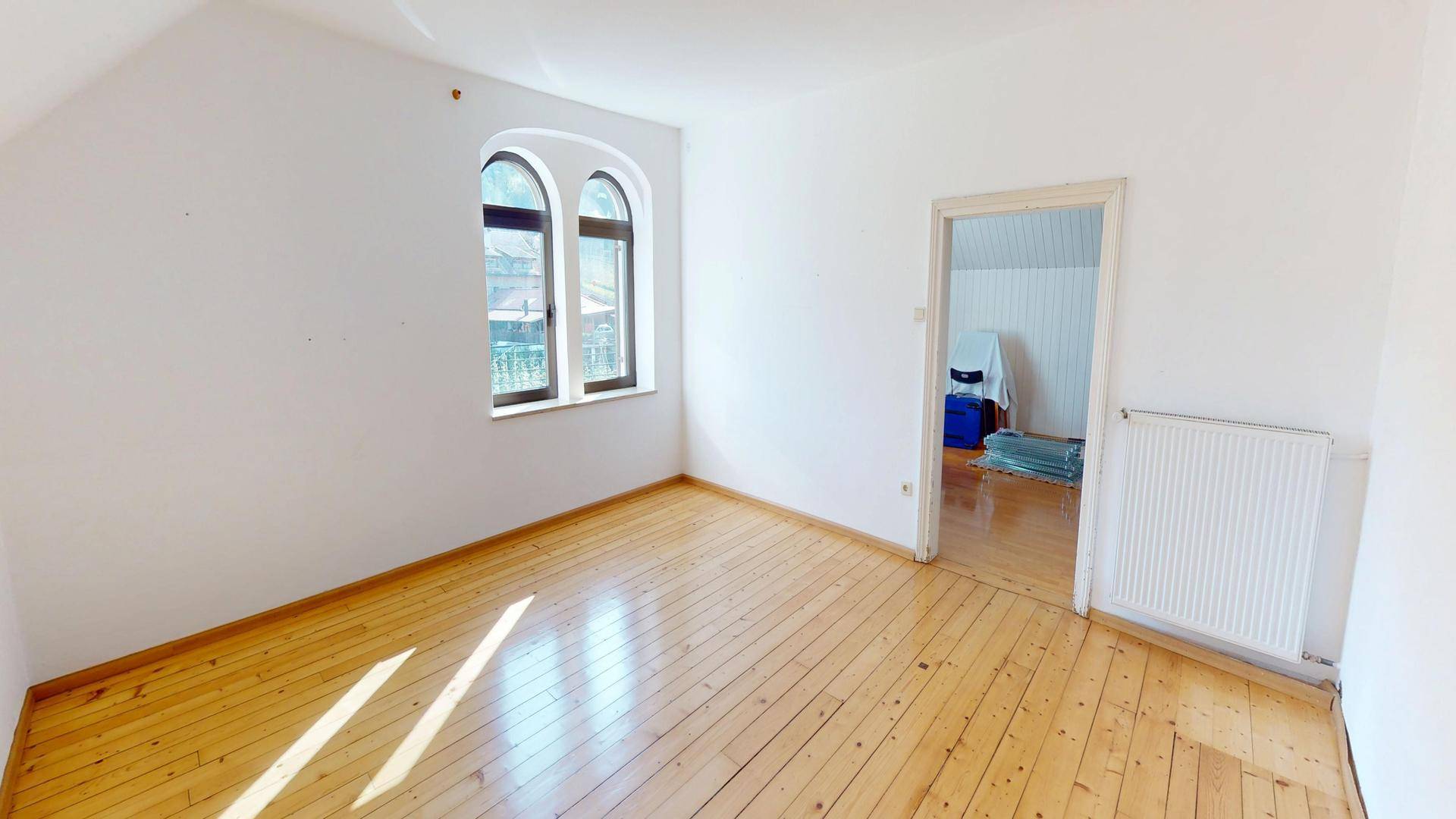 Helles Zimmer mit Holzdielenboden, Rundbogenfenstern und Zugang zu einem weiteren Raum.