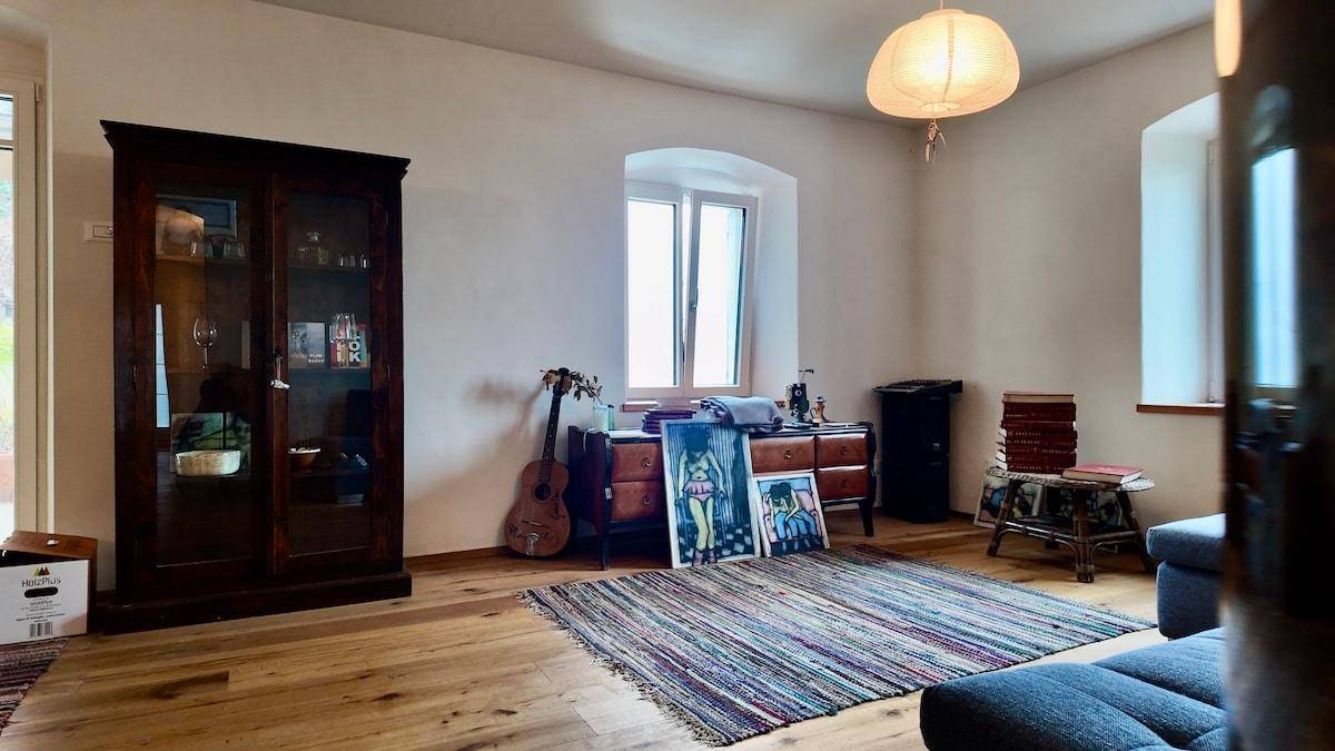 Helles Wohnzimmer mit Holzboden, großem Fenster und einem antiken Vitrinenschrank.