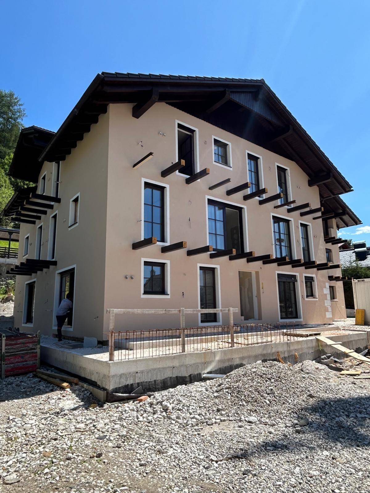 Ansicht des Neubaus von vorne und der Seite, mit beiger Fassade und großen Fenstern, umgeben von der Baustelle.