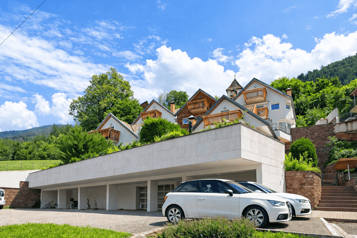 Wohnanlage mit Tiefgaragenzufahrt, geparkten Autos und grüner Hügellandschaft im Hintergrund.